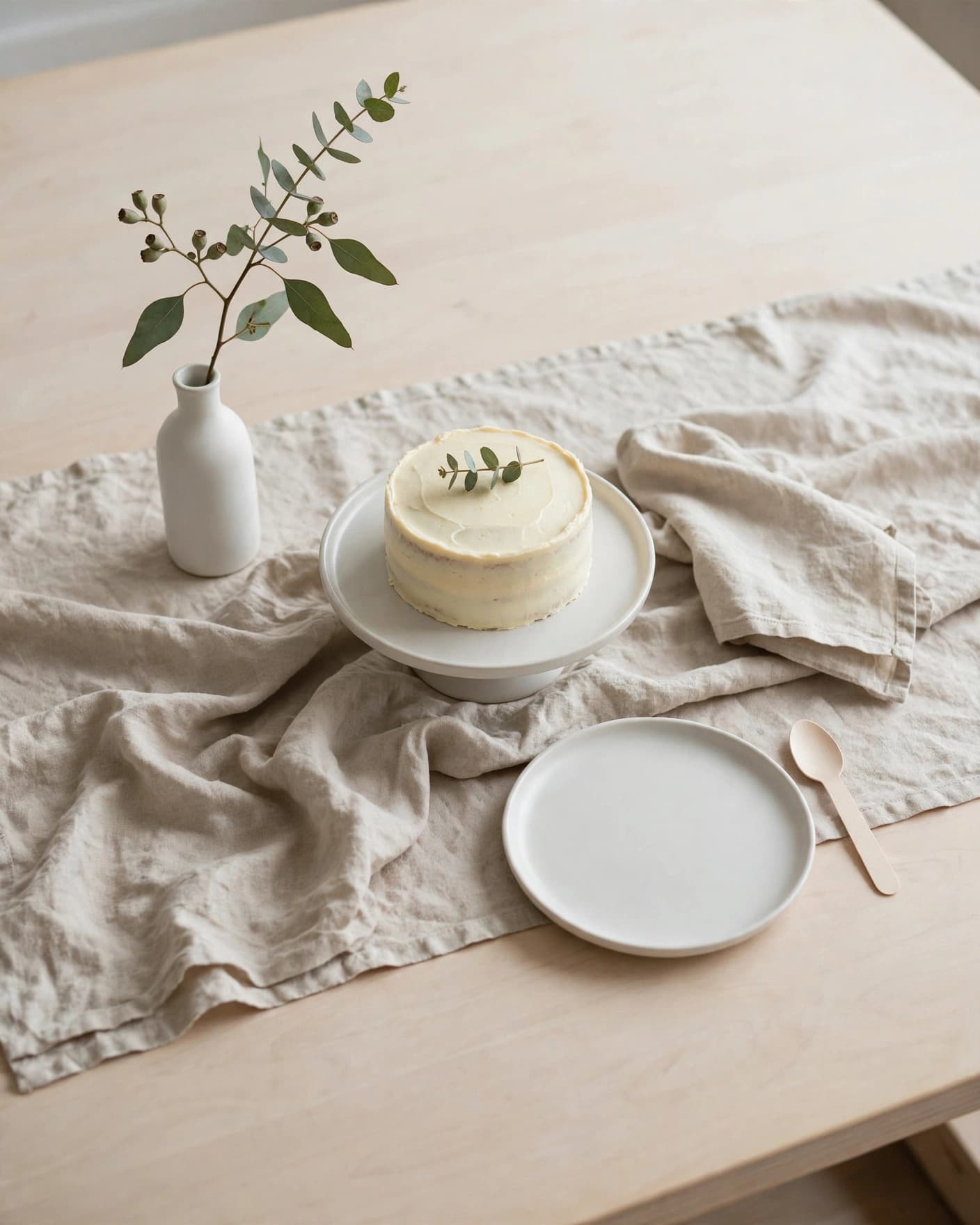 Minimalist baby sprinkle table with a small cake, white ceramics, beige linen, and a branch in a vase.